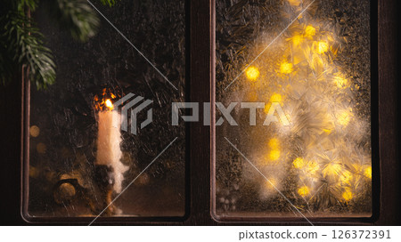Burning candle sits on a windowsill next to a frosted window pane with a christmas tree made of string lights glowing behind it during a cold winter night 126372391