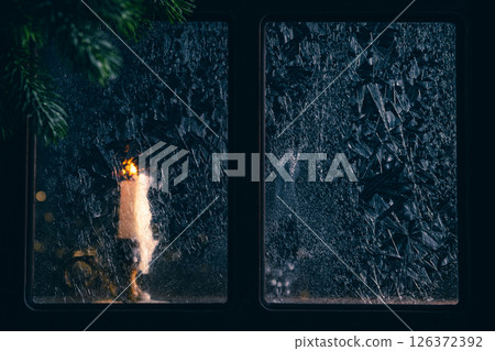 Single white candle is burning behind a frozen window during a cold winter night, creating a warm and cozy atmosphere with the fir tree branch in the foreground 126372392