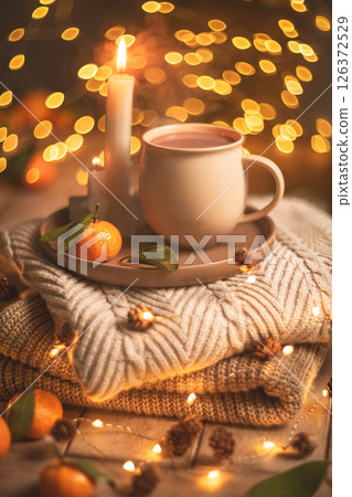Steaming cup of hot beverage sits on a tray with a mandarin orange and lit candles, placed on a cozy knitted blanket with warm, inviting lighting, creating a perfect winter atmosphere Steaming cup of hot beverage sits on a tray with a mandarin orange and lit candles, placed on a cozy knitted blanket with warm, inviting lighting, creating a perfect winter atmosphere 126372529