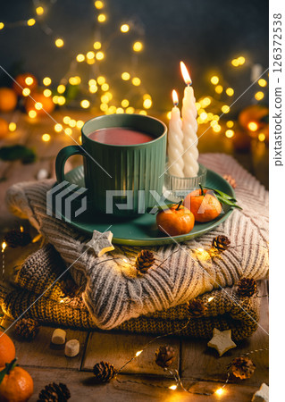 Green mug of hot chocolate resting on a plate with tangerines, surrounded by lit candles, warm sweaters, pine cones, and the soft glow of christmas lights, creating a cozy atmosphere, greeting card 126372538
