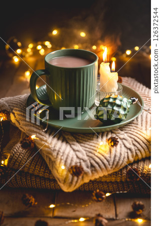 Christmas still life with mug of hot chocolate, candles and christmas lights on stack of knitted sweaters, creating cozy and festive atmosphere for christmas and new year celebrations, greeting card 126372544