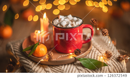 Red mug filled with hot chocolate and marshmallows resting on a tray with tangerines, surrounded by warm knit fabric and glowing christmas lights, creating a cozy winter scene Red mug filled with hot chocolate and marshmallows resting on a tray with tangerines, surrounded by warm knit fabric and glowing christmas lights, creating a cozy winter scene 126372549