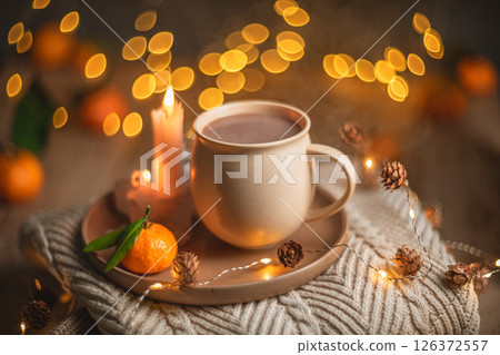 Christmas hot chocolate with tangerines and burning candles on a stack of knitted sweaters creates a cozy winter atmosphere with warm bokeh lights 126372557