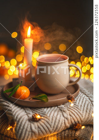 Steaming cup of hot beverage sits on a tray with a mandarin orange and lit candles, placed on a cozy knitted blanket with warm, inviting lighting, creating a perfect winter atmosphere 126372558