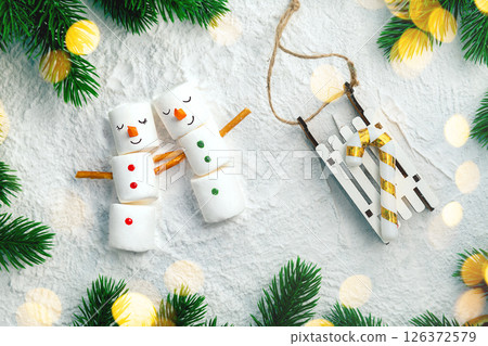Funny marshmallow snowmen holding hands lying on snow near christmas tree branches and a wooden sleigh with a candy cane are creating a festive atmosphere for christmas and new year holidays 126372579