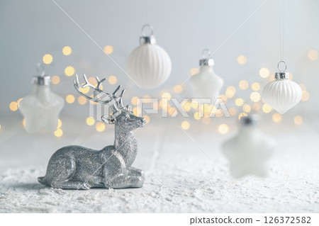 Silver reindeer figurine resting on snow-covered surface, surrounded by white christmas ornaments hanging amidst a backdrop of warm fairy lights, creating a magical and festive holiday atmosphere Silver reindeer figurine resting on snow-covered surface, surrounded by white christmas ornaments hanging amidst a backdrop of warm fairy lights, creating a magical and festive holiday atmosphere 126372582