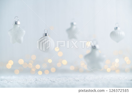 White christmas ornaments of various shapes hanging against a light gray background with soft, warm bokeh from sparkling lights, evoking a serene and festive holiday mood 126372585