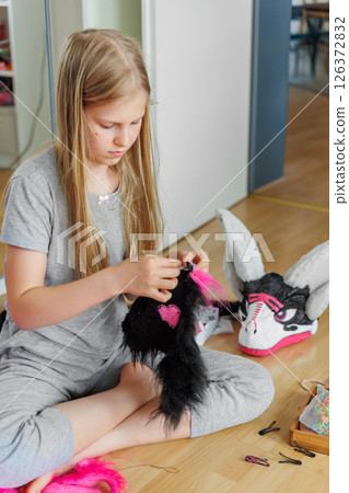 Cute teenager girl blonde hair sits floor sewing pink black fur costume piece. Child making homemade furry dragon mask. Quadrobic Therian fandom subculture. Design hobby creative artwork 126372832