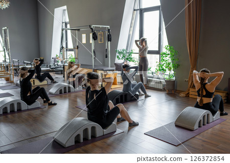 Group of women doing exercises on back spine corrector. Reformer pilates studio machine for fitness workouts in gym. Fit, healthy and strong authentical body. Fitness concept. Group of women doing exercises on back spine corrector. Reformer pilates studio machine for fitness workouts in gym. Fit, healthy and strong authentical body. Fitness concept. 126372854