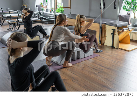 Group of women doing exercises on back spine corrector. Reformer pilates studio machine for fitness workouts in gym. Fit, healthy and strong authentical body. Fitness concept. Group of women doing exercises on back spine corrector. Reformer pilates studio machine for fitness workouts in gym. Fit, healthy and strong authentical body. Fitness concept. 126372875