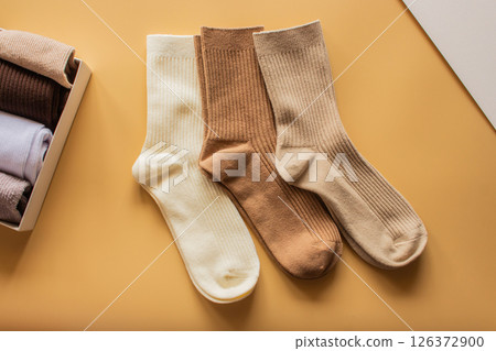 Three pairs of ribbed socks in cream, brown, and beige colors are arranged on warm yellow background, highlighting their cozy texture and stylish design. 126372900
