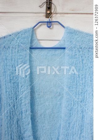 Blue knitted cardigan on hanger on white background. Soft blue knitted sweater in details. Knitting pattern background. Summer knitted fashion. Cozy hobby. Craft product concept.  126372989
