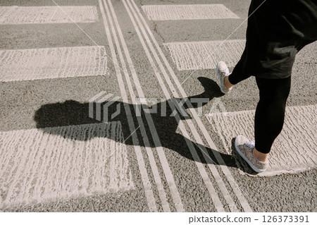 Woman walking across a pedestrian crosswalk with a clear shadow cast on the pavement. Perfect for Woman walking across a pedestrian crosswalk with a clear shadow cast on the pavement. Perfect for 126373391