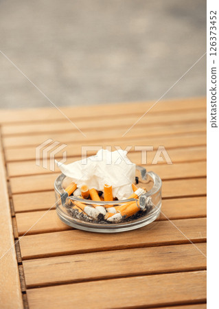 A glass ashtray filled with cigarette butts and tissue on a wooden cafe table. A detailed close-up 126373452