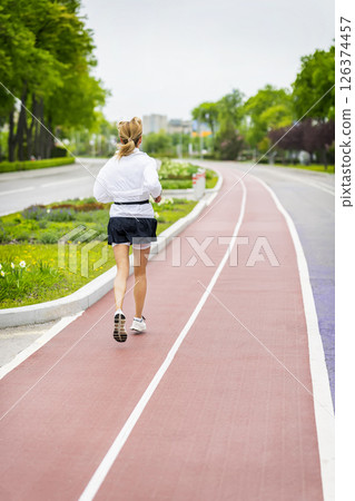Woman in white jacket running on a red track, urban stadium. Concept of jogging, active lifestyle, personal health, outdoor training motivation Woman in white jacket running on a red track, urban stadium. Concept of jogging, active lifestyle, personal health, outdoor training motivation 126374457