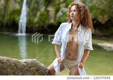Woman in lace panties and unbuttoned shirt posing by waterfall 126374948