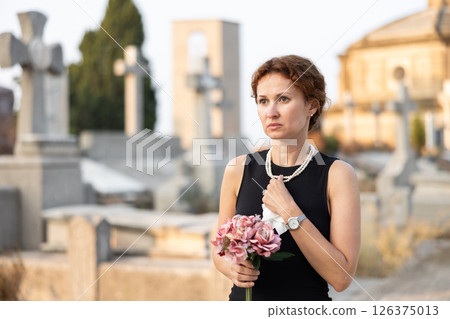 Grieving woman in black with flowers standing in cemetery 126375013