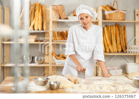 Woman works in bakery as baker, cuts dough into portions, forms pieces of dough to create buns. 126375067