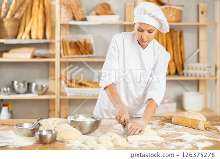 Female baker preparing croissant dough - kneading, rolling and cutting dough on table in bakery 126375278