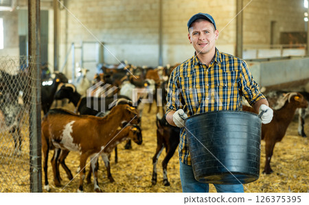 Portrait of male farmer standing with bucket and working in goat farmhouse. Feeding domestic animals with healthy organic food 126375395