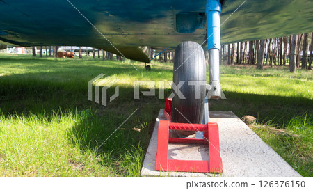 An old Soviet aircraft rests on a grassy area, supported by a red block beneath its tire, enveloped by lush trees, reflecting a piece of aviation history. 126376150
