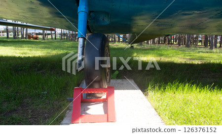 Beneath a vintage Soviet plane, a wheel rests on a colorful block amid lush grass and tall trees, evoking the nostalgia of Russian aviation. Beneath a vintage Soviet plane, a wheel rests on a colorful block amid lush grass and tall trees, evoking the nostalgia of Russian aviation. 126376152