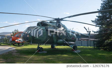A Soviet-era helicopter rests on display in an open area, showcasing its military design and vintage aesthetic against a picturesque backdrop of greenery and buildings. 126376176