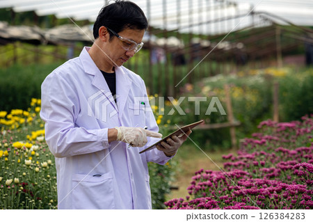 Scientist in white lab coat using digital tablet while inspecting blooming flowers in greenhouse field, wearing gloves and protective glasses, research vibrant floral surroundings in outdoor 126384283