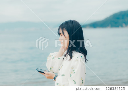 一位年輕女子在平靜的海面背景下操作智慧型手機 126384525