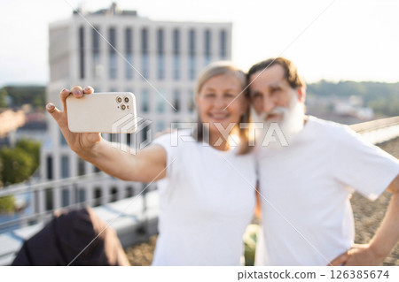 Elderly man with beard and elderly woman taking selfie outdoors in urban setting during sunny day, wearing casual white shirts, expressing joy and togetherness, enjoying outdoors, rooftop background 126385674