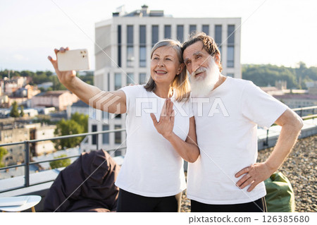 Elderly couple standing together on rooftop taking selfie during bright sunny day showcasing joy and companionship 126385680