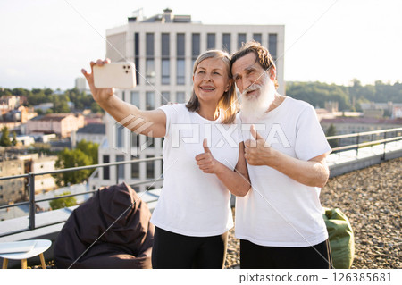 Elderly man and woman smiling while taking selfie outdoors, showing thumbs up, promoting active lifestyle and positivity in golden years. Elderly man and woman smiling while taking selfie outdoors, showing thumbs up, promoting active lifestyle and positivity in golden years. 126385681
