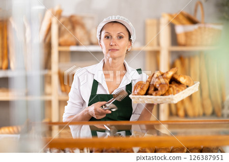 Selling fresh croissants in bakery - female baker places hot croissants on display 126387951