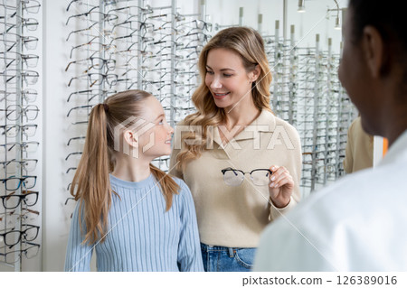 Mom and daughter choosing new eyeglasses in optic shop and looking interested 126389016