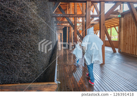 People wearing rain ponchos walk along a wooden spa walkway beside a brine graduation tower. The scene captures a relaxed, exploratory atmosphere in a unique architectural setting. 126391418