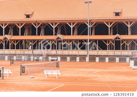 An outdoor clay tennis court is set against the backdrop of a large wooden structure. The court is empty, featuring benches, a net, and a ladder, conveying a peaceful setting 126391419