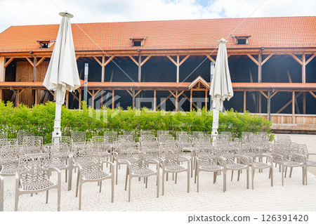 Rows of empty chairs with intricate designs are arranged outdoors in front of a large rustic wooden barn. Umbrellas are closed, and the setting is quiet, awaiting an event or gathering. Rows of empty chairs with intricate designs are arranged outdoors in front of a large rustic wooden barn. Umbrellas are closed, and the setting is quiet, awaiting an event or gathering. 126391420