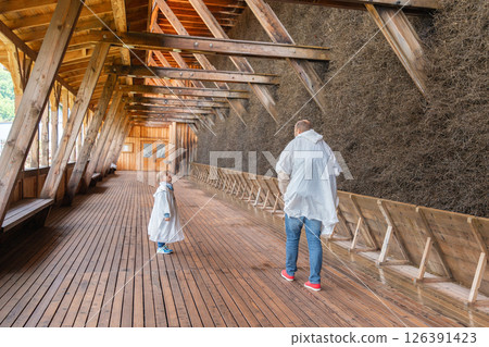 A father and son walk through a wooden corridor wearing raincoats. The child looks upward with curiosity, while the father explores the surrounding structure. A father and son walk through a wooden corridor wearing raincoats. The child looks upward with curiosity, while the father explores the surrounding structure. 126391423
