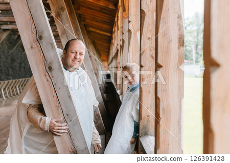 Father and young son wearing white protective coveralls, smiling on a wooden covered bridge. The setting suggests a serene outdoor environment, emphasizing their joyful bond. Father and young son wearing white protective coveralls, smiling on a wooden covered bridge. The setting suggests a serene outdoor environment, emphasizing their joyful bond. 126391428