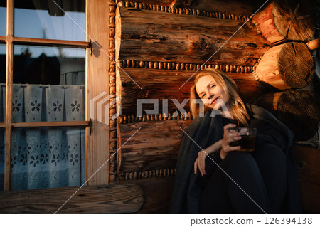 Smiling woman wrapped in blanket enjoying cup of hot beverage while sitting against log wall in cozy mountain chalet at sunset, creating peaceful and idyllic winter getaway scene. Woman sipping drink Smiling woman wrapped in blanket enjoying cup of hot beverage while sitting against log wall in cozy mountain chalet at sunset, creating peaceful and idyllic winter getaway scene. Woman sipping drink 126394138