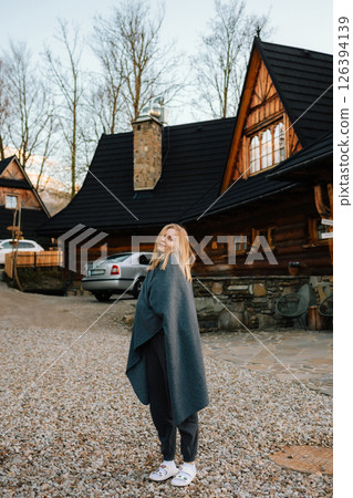 Blonde woman wearing gray blanket standing in driveway of her wooden chalet in mountains, enjoying fresh air and tranquility of nature. Mountain chalet wooden backdrop, woman wrapped in gray blanket 126394139