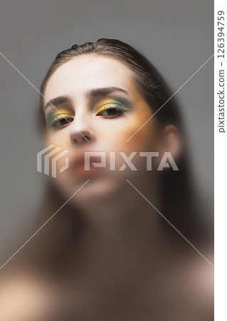 Artistic portrait. Woman with bold yellow and green eye makeup stares forward through soft diffusion, creating tension between intensity and haze. Artistic portrait. Woman with bold yellow and green eye makeup stares forward through soft diffusion, creating tension between intensity and haze. 126394759