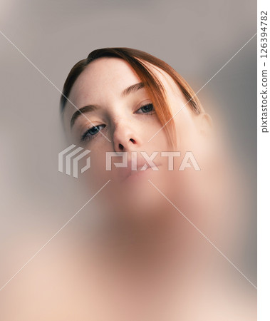 Conceptual artwork. Portrait of red-haired woman with piercing blue eyes looking downwards through blurred filter, conveying isolation and introspection. Conceptual artwork. Portrait of red-haired woman with piercing blue eyes looking downwards through blurred filter, conveying isolation and introspection. 126394782