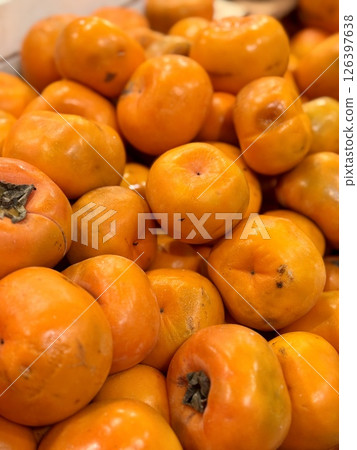 A pile of ripe juicy orange flat persimmons in a box, offered for sale at a supermarket vegetable stand, demonstrates organic, vegetarian and healthy food. Close-up. 126397638