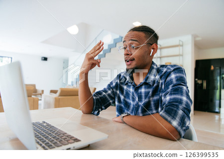 African American influencer in glasses sitting at a table in a modern living room, using a laptop for business video chat, conversation with friends and entertainment. African American influencer in glasses sitting at a table in a modern living room, using a laptop for business video chat, conversation with friends and entertainment. 126399535