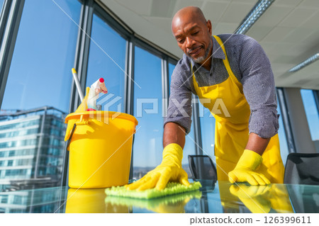 Mature senior man cleaning office glass desk with gloves and bucket in high-rise building 126399611
