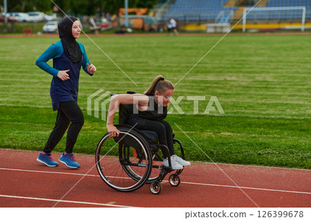 A Muslim woman in a burqa running together with a woman in a wheelchair on the marathon course, preparing for future competitions. 126399678
