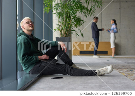 A casual young professional sitting by a large window in a modern office, smiling while working on a laptop, with colleagues interacting in the blurred background. 126399700