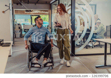 Young business colleagues, collaborative business colleagues, including a person in a wheelchair, walk past a modern glass office corridor, illustrating diversity, teamwork and empowerment in the 126399706