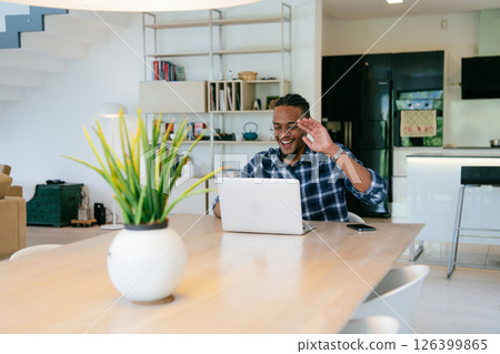 African American influencer in glasses sitting at a table in a modern living room, using a laptop for business video chat, conversation with friends and entertainment. 126399865